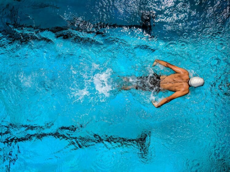 Top view of a swimmer wearing a cap, performing a front crawl stroke in a clear blue swimming pool.