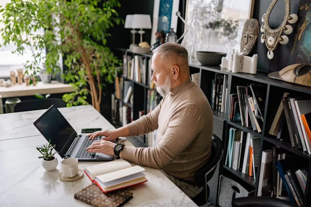 Senior man focused on laptop in a stylish home office, surrounded by books and plants.