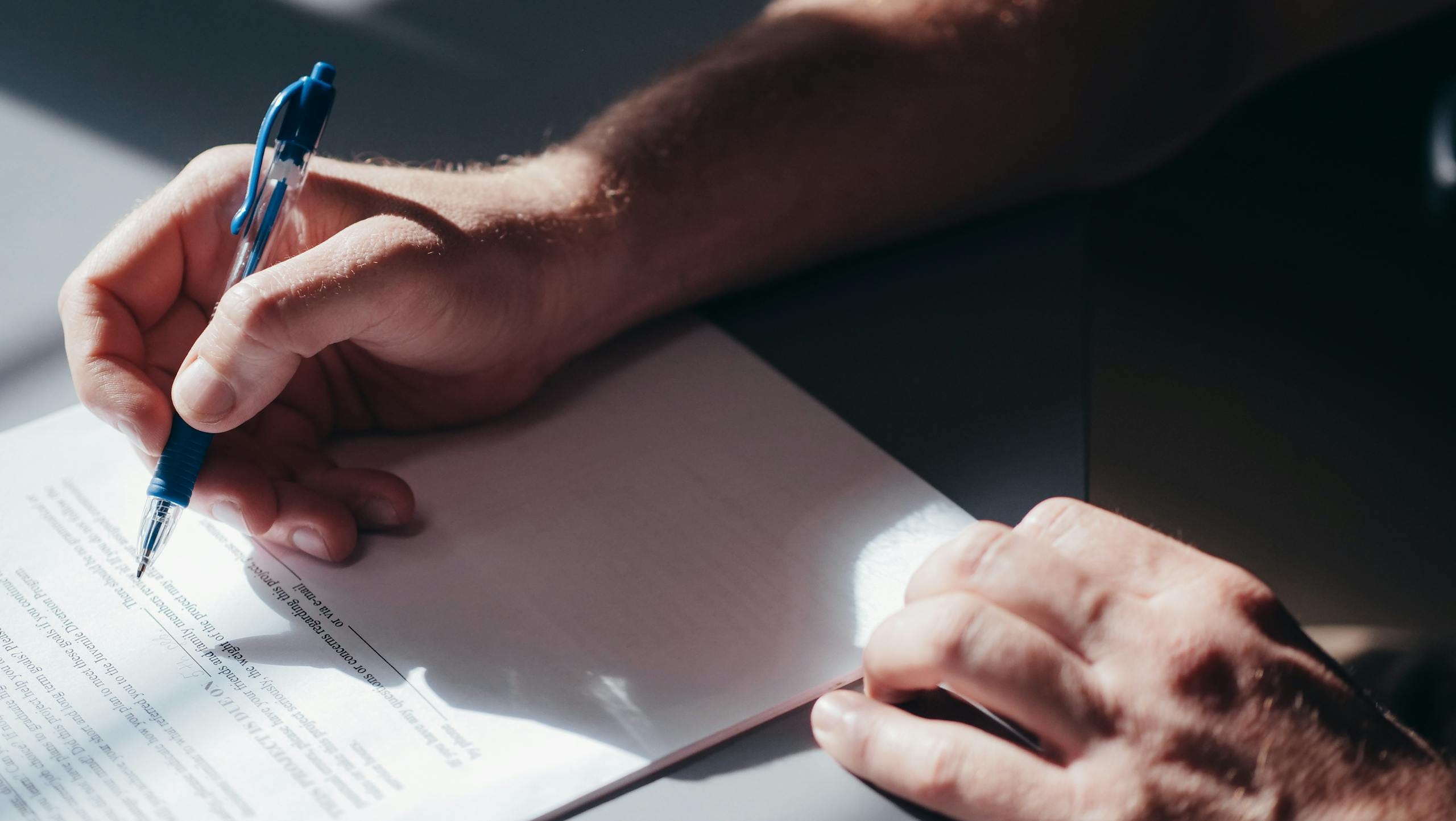 A close-up image of a person's hand signing a document with a blue pen in natural lighting.