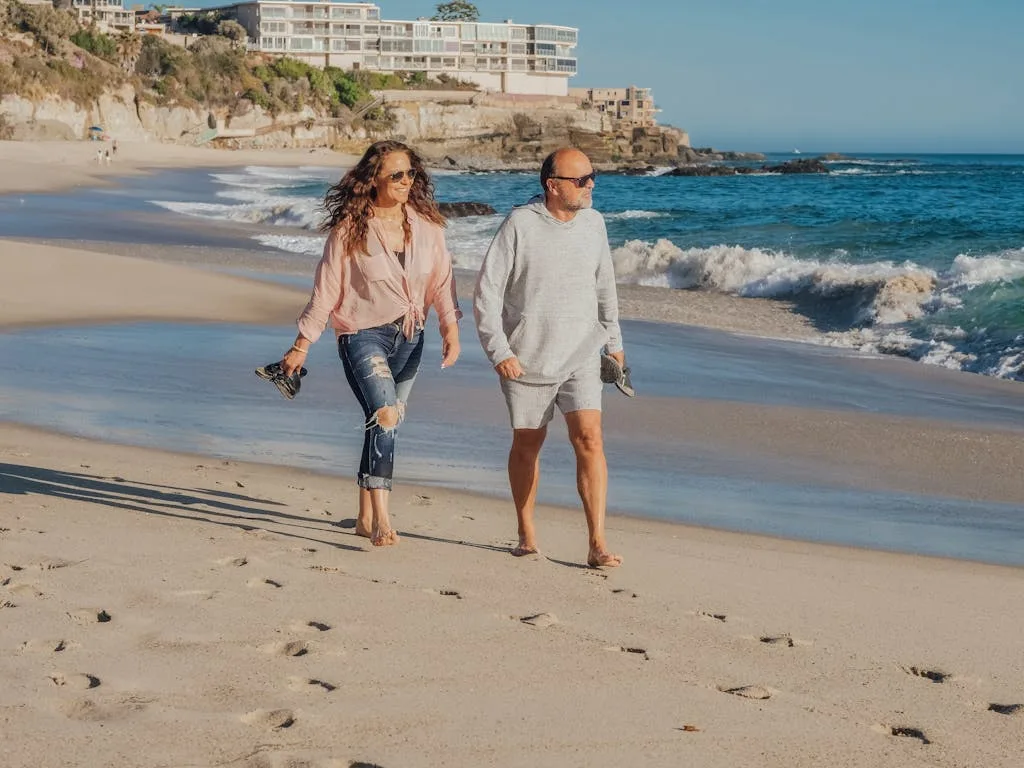 Elderly couple walking barefoot on a sunny beach by the coast, enjoying summer vacation.
