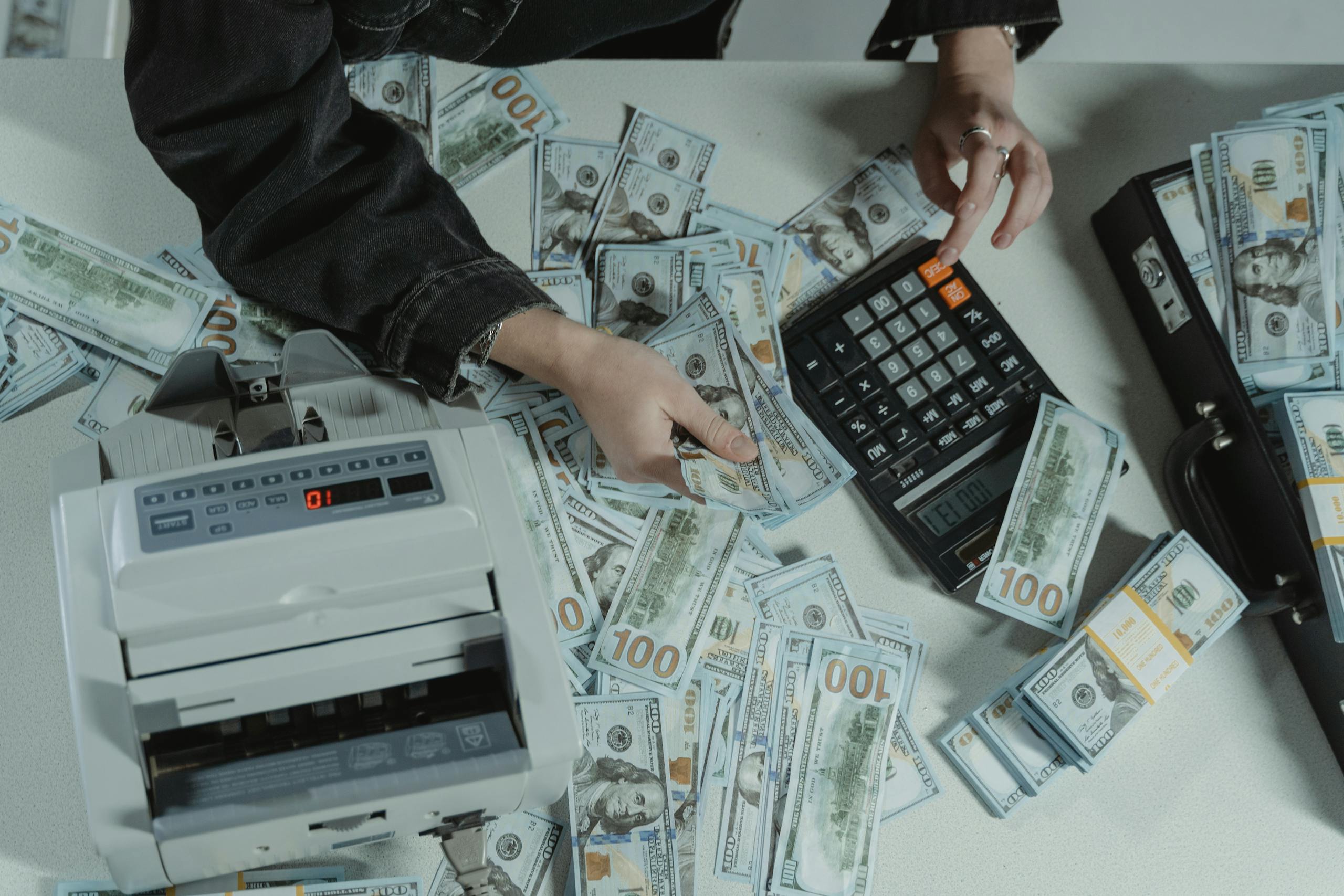 Hands counting 100 dollar bills using a calculator and money counter on a table.